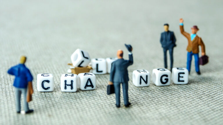 Business figurines surrounding letter blocks spelling “CHANGE,” with one letter fallen, symbolising how teams recognise the need for change but struggle to act on it.